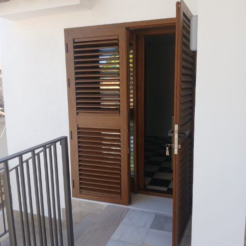 Puerta exterior de aluminio con contraventana y lamas en acabado imitación madera para vivienda unifamiliar.